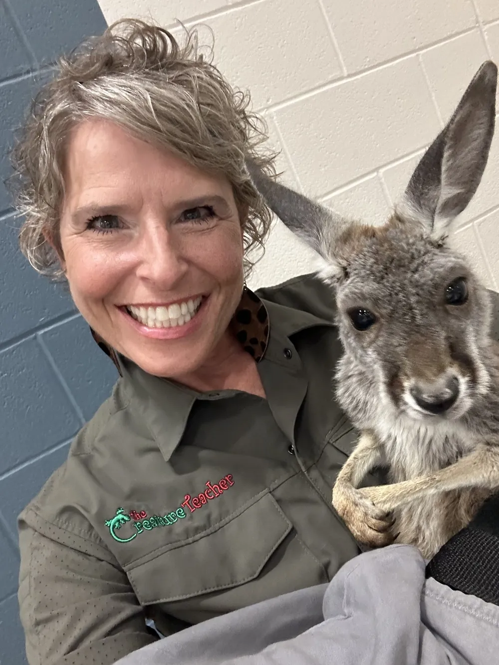 A wildlife educator smiles beside a young kangaroo held in a carrying pouch.