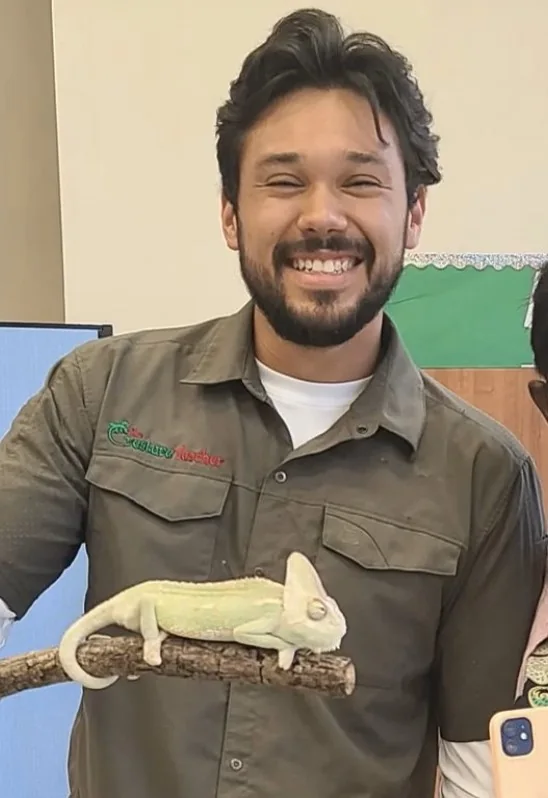 An educator smiles while a pale green chameleon perches on a branch he is holding.